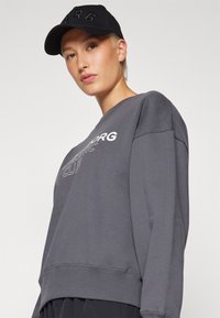 Sudadera gris oscuro con cuello redondo y letra bordada en blanco. Combinada con una gorra negra con texto en relieve. Textura suave.