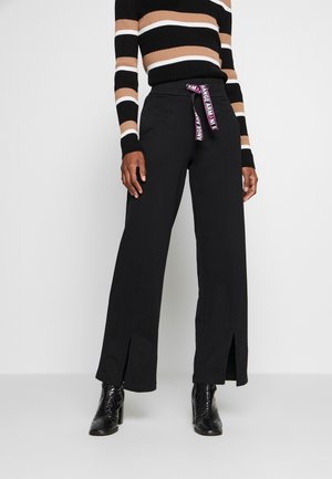 Personne portant un pantalon noir à jambes larges avec des fentes à l'avant et une ceinture rose et blanche marquée à la taille, un haut à manches longues rayé et des bottines noires à talons.