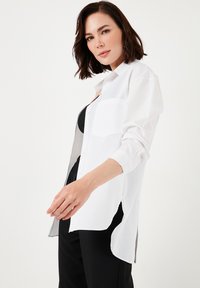 White button-up shirt made of lightweight fabric, featuring a breast pocket, side slits, and long sleeves, worn over a black top and pants.