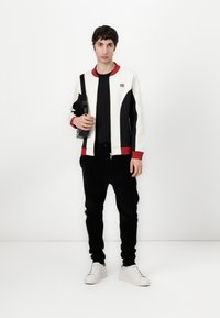 Herre zip-up bomberjakke i kremfarge, svart og rød; har et fargeblokkdesign med ribbede mansjetter. svarte corduroy bukser og hvite sneakers.