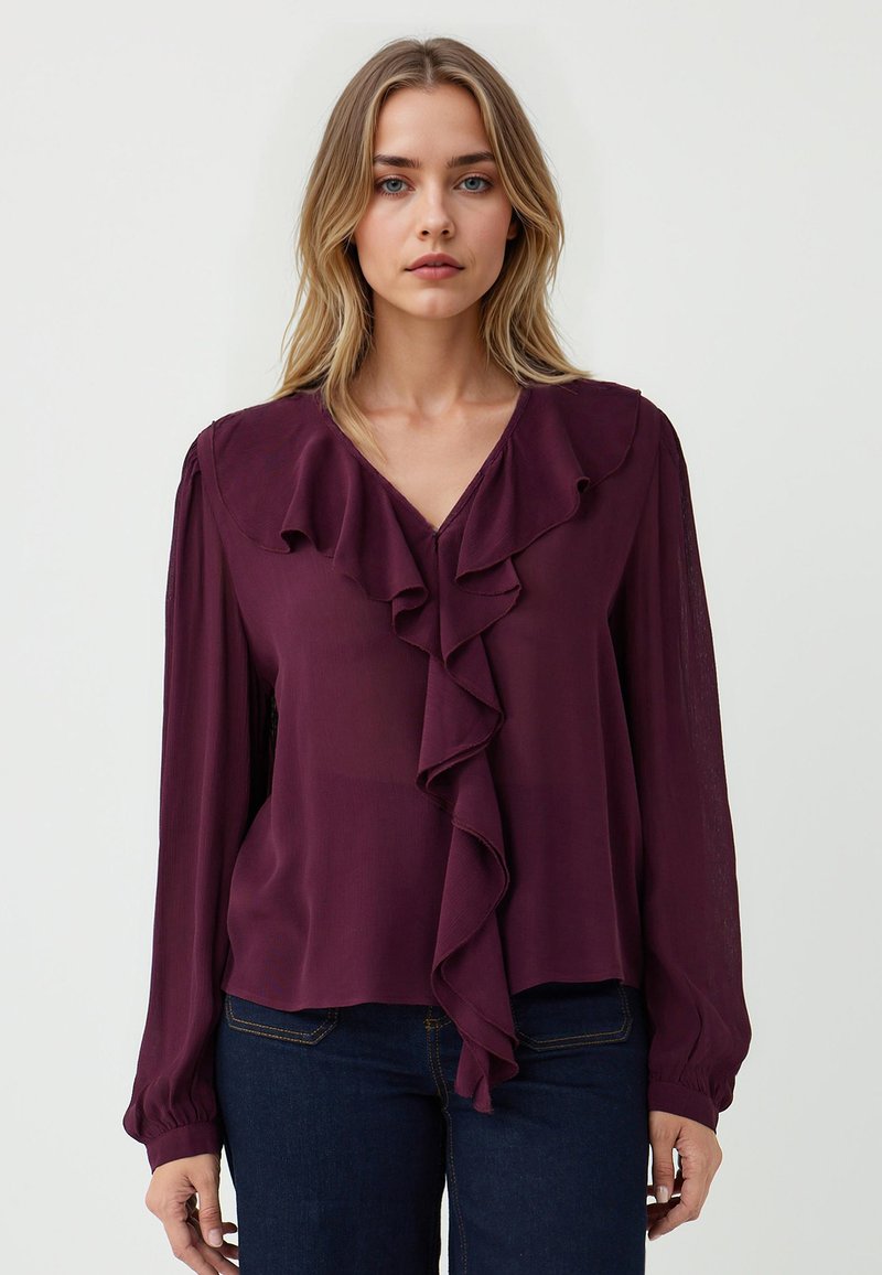 Blusa viola realizzata in tessuto leggero, con scollatura a V, maniche lunghe e un dettaglio a volant sul davanti. Abbinata a jeans blu scuro.