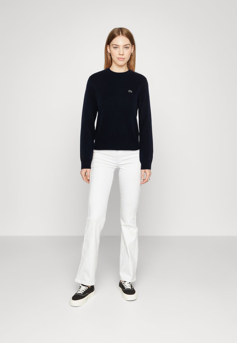 Maglione in maglia blu navy con scollo a giro e orlo a costine, abbinato a jeans bianchi a zampa d'elefante e sneakers nere con suole bianche. Accento del logo orizzontale.