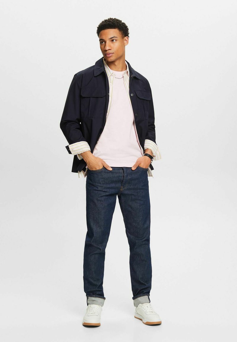 Marineblaues Jackett mit zwei Vorderseitentaschen, pinkes T-Shirt darunter, dunkelblaue Jeans und weiße Sneakers mit braunen Sohlen. Minimalistisches Design und Texturen.