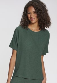 Jauna sieviete ar cirtainiem matiem, valkājot vaļēju, ribotu tumši zaļu īsroku t-kreklu, nedaudz smaida uz vienkārša balta fona.