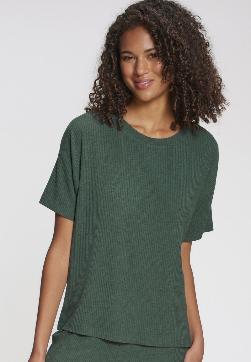 Jauna sieviete ar cirtainiem matiem, valkājot vaļēju, ribotu tumši zaļu īsroku t-kreklu, nedaudz smaida uz vienkārša balta fona.