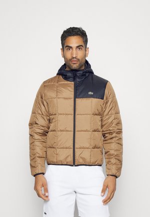 Chaqueta acolchada marrón y negra con cremallera frontal, capucha fija y puños elásticos. Presenta un pequeño logo en el pecho. Material suave y ligero.