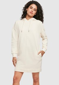 Vestito con cappuccio color crema realizzato in tessuto morbido. Presenta una tasca frontale, maniche lunghe e coulisse regolabili sul cappuccio.