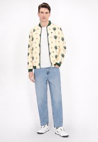 Bomberjack met palmboom- en kokosnootafdruk, crème stof, groene geribbelde kraag en zoom, gecombineerd met lichtblauwe jeans en witte sneakers.
