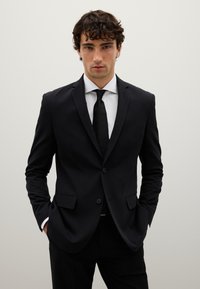 Completo sartoriale nero con vestibilità slim, caratterizzato da rever a punta, due bottoni e due tasche frontali. Abbinato a una camicia bianca e una cravatta nera.