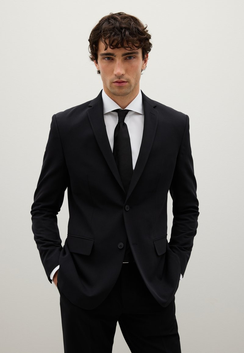 Completo sartoriale nero con vestibilità slim, caratterizzato da rever a punta, due bottoni e due tasche frontali. Abbinato a una camicia bianca e una cravatta nera.