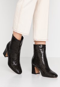 Botas negras de tobillo con una textura de serpiente brillante, puntera afilada y tacón grueso. Acentuadas con un ligero corte en el costado.