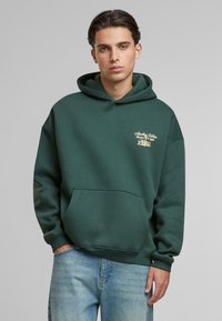 Dark green hoodie made of soft fabric, featuring a front pocket, and gold logo detailing on the chest. Relaxed fit with a drawstring hood.