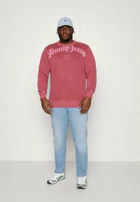 Un uomo indossa un felpa rossa "Tommy Jeans", jeans azzurri chiari, sneakers bianche e un cappellino azzurro chiaro, in piedi su uno sfondo neutro.