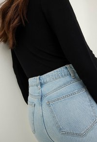 Jeans denim biru muda dengan pinggang tinggi, dilengkapi dengan sabuk bermerek, kantong belakang, dan tekstur yang halus. Dipadukan dengan atasan lengan panjang hitam.