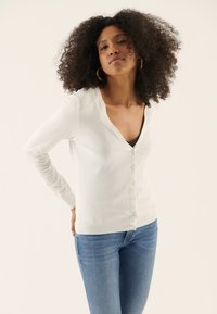 White, long-sleeve cardigan with a V-neck, featuring a button-down front and soft texture. Worn over a black top, paired with blue jeans.