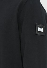 Sort sweatshirt med rund halsudskæring og lange ærmer, lavet af blødt materiale, med et kvadratisk vævet 'Weekend Offender'-mærke på ærmet.