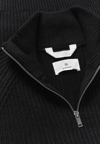 Suéter negro con textura acanalada, cuello alto y cremallera en la parte delantera. La etiqueta interior dice "Matinique" en negro sobre un fondo blanco.