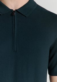 Donkergroen gebreide polo met een ritssluiting bij de kraag. Korte mouwen en een gladde textuur benadrukken het moderne ontwerp.