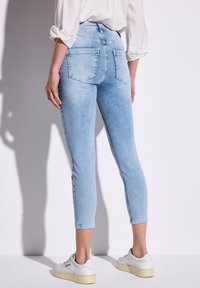 Pantalons capri en denim bleu clair au design ajusté, avec deux poches arrière et un ourlet légèrement effiloché, associés à des baskets blanches.