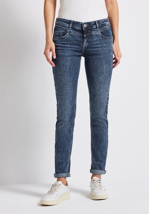 Woman wearing dark blue slim-fit jeans paired with white sneakers and a white top, standing against a plain light background.