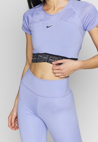 Kvinna som bär en lavendel Nike Pro sporttopp med meshdetaljer och matchande högmidjade leggings, som lyfter toppen något för att avslöja midjan.
