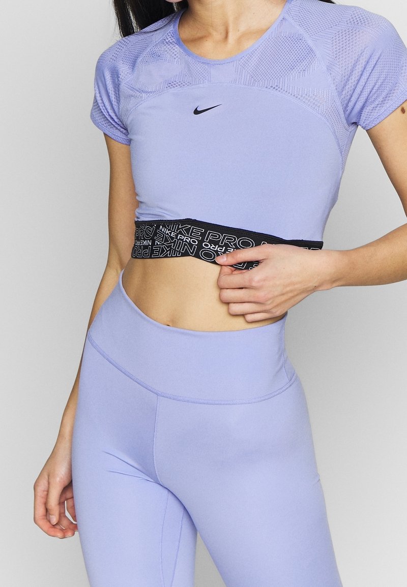 Kvinna som bär en lavendel Nike Pro sporttopp med meshdetaljer och matchande högmidjade leggings, som lyfter toppen något för att avslöja midjan.