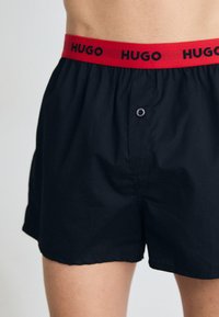 Boxer shorts en coton noir avec une ceinture élastique rouge portant l'inscription "HUGO" et un seul bouton à l'avant pour la fermeture.