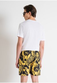 Camiseta blanca de cuello redondo combinada con pantalones cortos de baño amarillos con patrones de hojas negras. Modelo visto desde atrás, llevando gafas de sol oscuras.