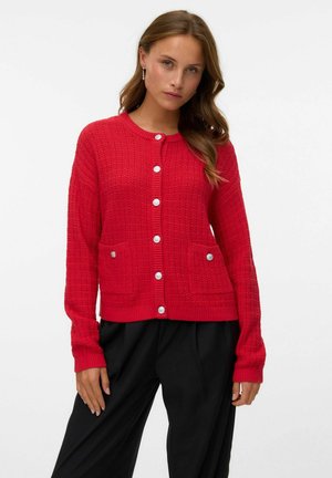 Vrouw in een rode, gestructureerde cardigan met knopen en een paar voorzakken, gekleed in zwarte, wijde broek, staand voor een effen witte achtergrond.