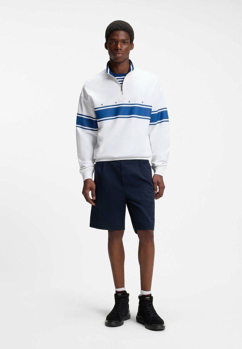 Homme debout portant un sweat zippé blanc avec des rayures bleues, un short bleu marine, des chaussettes blanches et des baskets montantes noires sur fond blanc.