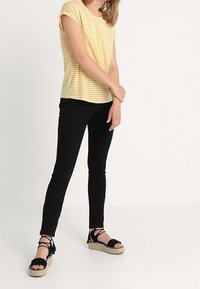 Żółto-biało paskowana koszula z krótkim rękawem, czarne wąskie dżinsy oraz czarne espadryle z wiązaniem. Model stoi w neutralnym otoczeniu.