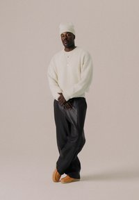 Cremefarvet, strikket sweater; løse, sorte læderbukser; lyse slippers; lagdelte guldkæder; struktureret, strikket hue; neutral baggrund.