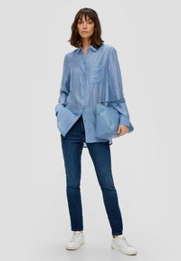 Helles blaues Hemd mit Knopfverschluss und Brusttasche, kombiniert mit dunkelblauen Jeans und weißen Sneakers. Weicher Stoff, lockere Passform, lange Ärmel.
