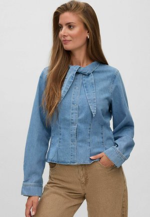 Kvinde med langt hår iført en lyseblå knappet denimskjorte med bundet krave og beige bukser, stående med den ene hånd i lommen.