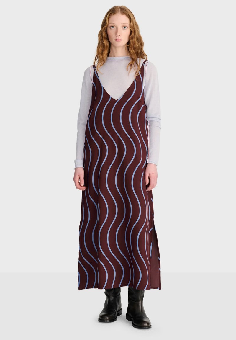 Femme aux cheveux ondulés porte une robe marron sans manches avec des lignes ondulées bleues sur un haut clair à manches longues et des bottes noires, debout face à l'avant.