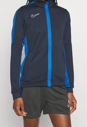 Personne portant une veste de sport zippée bleu marine et bleu avec un short noir à motifs, debout devant un fond clair uni.