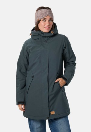 Une femme porte un manteau d’hiver à capuche bleu canard foncé et un bandeau en tricot mauve. Elle a une main dans une poche, sur un fond uni.
