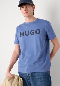 Mėlyna medvilninė marškinė su ryškiai juodu „HUGO“ užrašu, apvaliu kaklu, trumpomis rankovėmis ir laisvu siluetu, derinama su džinsais ir smėlio spalvos viršutiniu drabužiu.