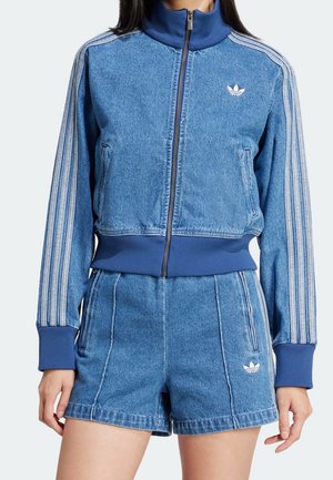 Vrouw draagt een blauwe denim Adidas trainingsjas en bijpassende shorts met witte strepen op de mouwen en Adidas-logo's op de borst en shorts.
