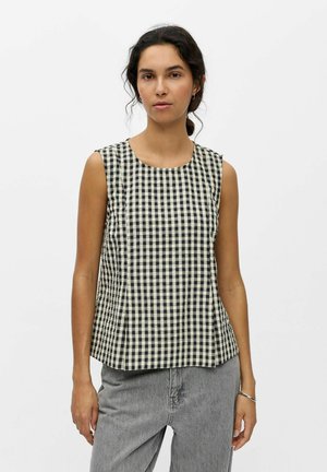 Žena s tmavými vlasmi v copu, ktorá má na sebe čierno-biely kockovaný tielko a sivé džínsy, stojaca pred bielym pozadím.