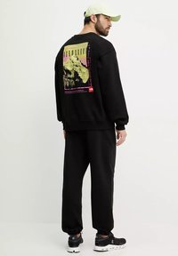 Sudadera negra con diseño gráfico que presenta una montaña y texto en la parte posterior. Combinada con pantalones de chándal negros y zapatillas negras. Gorra clara.