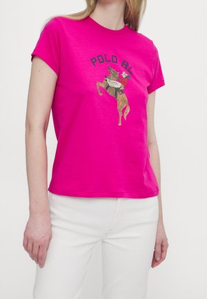 Vrouw draagt een felroze T-shirt met een afbeelding van een paard en ruiter en de tekst "POLO RL", gecombineerd met een witte broek.
