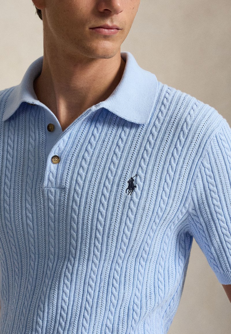 Jeune homme portant un polo en maille torsadée bleu clair à manches courtes avec un logo brodé foncé sur la poitrine, fond neutre.