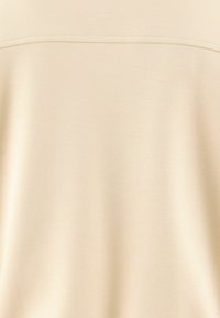 Top beige chiaro con una texture liscia e un design semplice. Presenta una cucitura orizzontale nella zona del petto, senza altri accenti visibili.