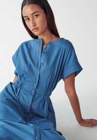 Gumbna denim obleka v svetlo modri barvi, ki ima zategnjeno pas, kratke rokave in ramenske šive s sproščenim krojem.