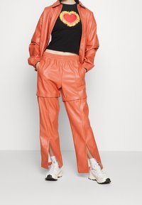 Coral faux leather jacket and pants set; pants feature snap details and slits. Black shirt with a colorful heart graphic; worn with white sneakers.