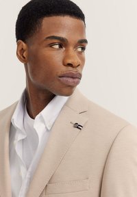 Beige Blazer aus strukturiertem Stoff, mit Reverskragen und einer Einstecktuch. Darunter ein weißes Hemd mit Knöpfen. Als Accessoire ein Anstecker mit Streifen.