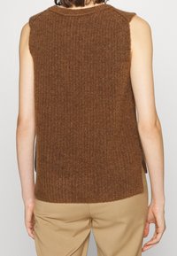 Chaleco de punto sin mangas color marrón con textura acanalada, cuello alto y aberturas laterales, fabricado en hilo suave. Llevado sobre pantalones beige.