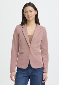 Blazer rosa texturizado con solapas con muesca, cierre de un botón y dos bolsillos delanteros. Combinado con una camiseta beige y jeans azul oscuro.
