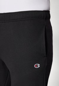 Pantalones de chándal negros de algodón con cintura elástica, bolsillos laterales y un pequeño logotipo bordado en el muslo. Textura suave, corte relajado.